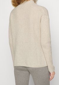 Personne portant un pull en tricot texturé de couleur crème et un pantalon en tricot côtelé gris, tourné le dos avec la main relâchée sur le côté.