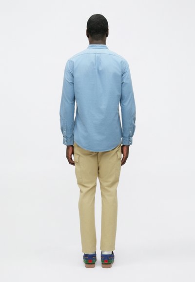 Camisa azul clara de manga comprida com punhos abotoados, combinada com calças cargo bege. O indivíduo usa calçado multicolorido, visto de costas.