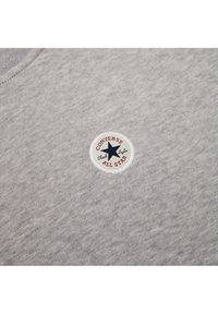 Sudadera gris con una tela que presenta un parche blanco redondo con una estrella azul y texto rojo que dice "Converse Chuck Taylor All Star."