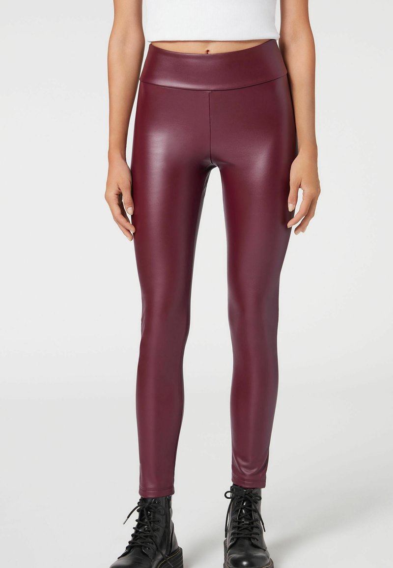 Leggings en simili cuir bourgogne avec taille haute, coupe près du corps et texture lisse, associés à des bottes noires à lacets.