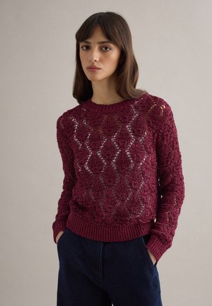 Donna che indossa un maglione a maglia aperta color bordeaux con motivi a diamante e pantaloni blu scuro, in piedi con le mani nelle tasche davanti a uno sfondo semplice.