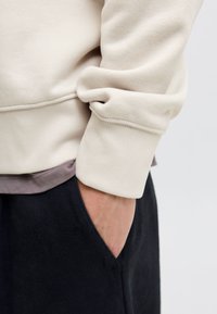 Osoba ubrana w beżową bluzę z mankietem, z ręką schowaną w kieszeni czarnych spodni na tle jednolitego jasnego tła.