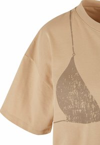 Beež t-särk, millel on laienev lõige, lühikesed varrukad ja graafiline print kergelt tumedamas beeži toonis, millel on tekstuurne välimus.