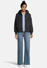 Veste bomber noire à fermeture éclair avec capuche, poignets et ourlet côtelés. Associée à un haut bleu clair et un jean en denim à jambes larges. Des baskets blanches complètent le look.