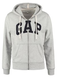 Felpa grigia con zip realizzata in misto cotone, con un grande logo "GAP" in blu navy. Presenta un cappuccio con cordino e due tasche frontali.