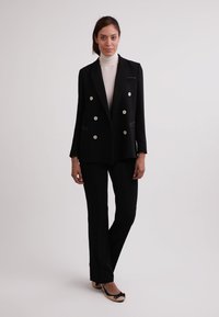 Černý dvouřadý blazer se stříbrnými knoflíky, klopami a přiléhavým střihem, v kombinaci s odpovídajícími černými kalhotami a plochými černými botami.