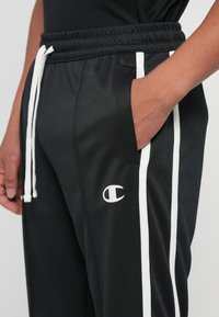 Person, der schwarze Sporthosen mit weißen Streifen, weißen Kordeln und Champion-Logo trägt, mit der rechten Hand in der Tasche.