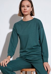 Tökéletes zöld, hosszú ujjú póló, kerek nyakkivágással, laza szabással és puha anyagból készült. Bordázott mandzsetta jellemzi, minimalista dizájnnal; összeillő nadrággal viselve.
