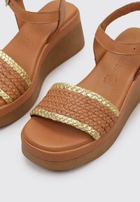 Tan wedge sandals with a braided, gold-accented strap. Smooth leather upper, cushioned footbed, and a sturdy, textured platform sole.