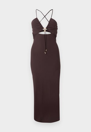 Kobieta stoi, jedną ręką opierając się na biodrze, a drugą na udzie, ubrana w ciemnobrązową sukienkę midi na szyję i czarne sandały na wysokim obcasie.