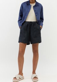 Chaqueta de lino azul marino con cuello y botones, combinada con pantalones cortos de lino negros con cintura elástica. Sandalias beige con tiras cruzadas.