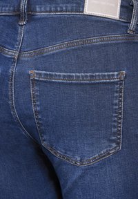 Jeans de mezclilla azul oscuro con un bolsillo trasero que presenta costuras en contraste y una etiqueta que dice "Street One Studio" en la cinturilla.