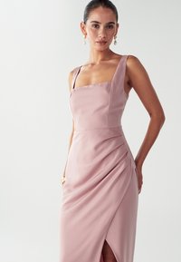 Moteris vilkinti berankovį rožinės satino suknelę su kvadratiniu iškirpimu ir šoninėmis klostėmis, accessorized su perlų auskarais ir auksiniu apyranke.