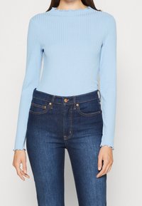 Helles blaues, geripptes Langarmoberteil mit gewelltem Kragen, kombiniert mit dunkelblauen, hoch taillierten Jeans mit goldenen Applikationen und Nähten.