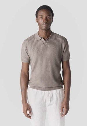 Fiatal férfi rövid göndör hajjal, taupe színű, texturált rövid ujjú polóban és fehér nadrágban, sima világos háttér előtt állva.