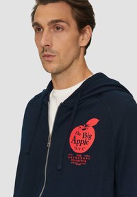 Felpa blu navy con cerniera frontale, caratterizzata da una grafica di una mela rossa e dalla scritta "The Big Apple N.Y.C." sul petto. Materiale in cotone, texture morbida.