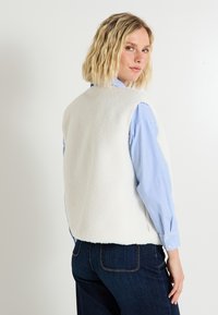 Gilet blanc doublé en polaire avec une texture douce, présentant un col rond, un design sans manches et un dos légèrement court. Porté par-dessus une chemise à rayures bleues.