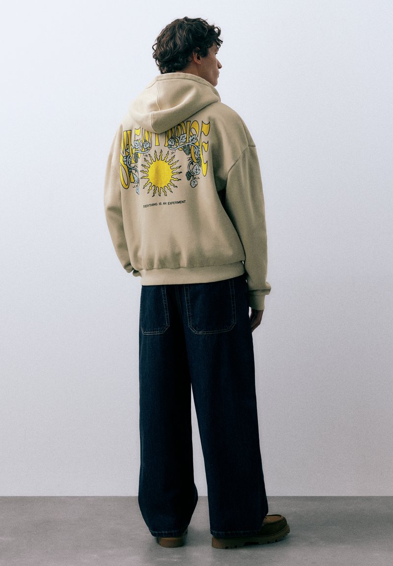 Mann trägt beige Kapuzenpullover mit Sonnenmotiv und dem Text „ALLES IST EIN EXPERIMENT“, dunkle weite Jeans und braune Schuhe, steht seitlich.