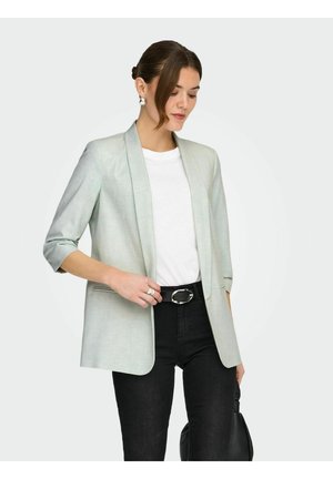 Vrouw die een lichtgrijze blazer draagt over een wit shirt, zwarte broek en een zwarte riem, en met één hand een zwarte tas vasthoudt.