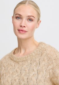 Raskaan beige neulepaita, jossa on teksturoidut palmikkokuvio, leveä pyöreä kaula-aukko ja pehmeät, pörröiset kuidut.