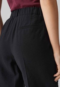 Pantaloni neri con elastico in vita, dotati di una tasca posteriore, dalla texture liscia e dal fit rilassato. Il tessuto ha una leggera lucentezza.