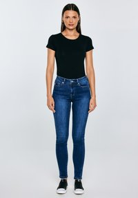 Vrouw staand, gekleed in een zwart T-shirt met korte mouwen, blauwe strakke jeans, zwarte sneakers, met steil donker haar tegen een effen lichte achtergrond.