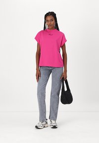 Pink short-sleeve T-shirt with a crew neck and embroidered logo, paired with gray jeans and a black woven handbag.