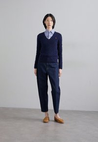 Jūros laivyno megztinis su pynėmis, derinamas su šviesiai mėlyna marškinėliais su apykakle ir tamsiai mėlynomis džinsinėmis kelnėmis. Rudos loafer kedai užbaigia aprangą. Neutralus fonas.