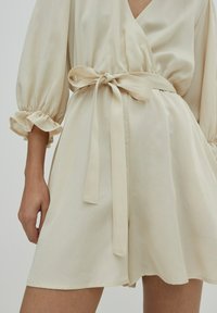 Vestido envolvente beige con mangas cortas abullonadas, atado en la cintura con un cinturón a juego. Tejido suave con un ligero brillo y falda acampanada.