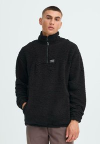 Schwarzer Fleece-Pullover mit hohem Kragen und halber Reißverschluss, mit einer aufgesetzten Tasche mit Logo und strukturierter, flauschiger Oberfläche.