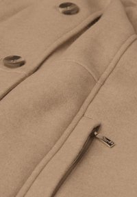 Beige wollen jas met grote knopen, een ingeknipt kraag en een zijrits met een metalen trekker. De stof heeft een zachte, textuurafwerking.