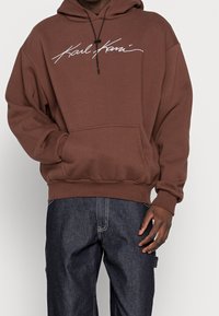 Brun hoodie med snörning i huvan och framficka. Har en vit "Karl Kani" logotyp över bröstet. Slät textur, avslappnad passform.