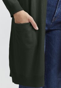 Donkergroene cardigan met geribde manchetten en een voorzak, met een gladde textuur. Gecombineerd met blauwe spijkerbroeken.