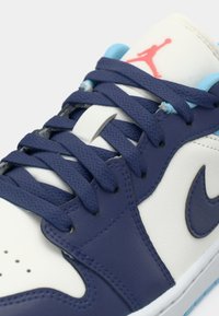Baltas sporta apavi ar tumši zili akcentiem, ar teksturētu ādas virsu, apaļo purngalu, tumši zilām šņorēm un rozā Jumpman logotipu uz mēles.