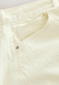 Gros plan sur la poche d'un pantalon ivoire montrant des coutures diagonales, un rivet et une ouverture courbe sur un tissu texturé.