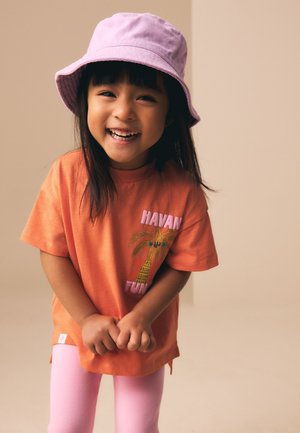Mladá dívka se usmívá, má na sobě růžový klobouk typu bucket, oranžové tričko s grafikou palmy a světle růžové kalhoty, stojící před béžovým pozadím.