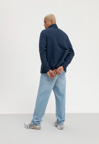 Jachetă din fleece de culoare bleu marin, cu guler înalt, purtată cu blugi lejeri de culoare bleu deschis și adidași atletici multicolori. Design minimalist.
