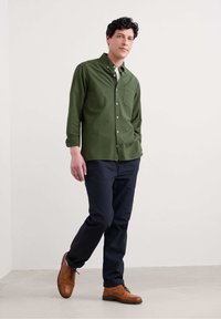Groene overhemddoek met een borstzak, gecombineerd met donkerblauwe broek en leren schoenen in bruin. Soepele stofstructuur met opgerolde mouwen.