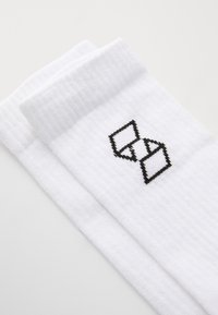 Fehér bordás zoknik, oldalán fekete geometriai logóval hímzve. A design egyszerű, puha tapintású.