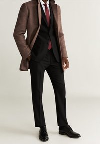Brązowy płaszcz z wełny na czarnym garniturze, białej koszuli i bordowym krawacie, w zestawieniu z czarnymi skórzanymi butami. Dopasowany krój, wyrazisty design.
