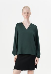 Grønn, langermet bluse med V-hals; laget av glatt stoff; løs passform med rynkede mansjetter; brukt over et svart skjørt.