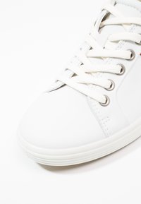 Primo piano della parte anteriore di una sneaker bianca con lacci bianchi infilati attraverso occhielli in metallo e dettagli puliti della suola in gomma.