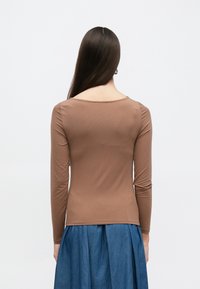 Pruun pikkade varrukatega ülikleeplib kangast topp, millel on kaarekujuline kaelus ja keha järgnev lõige; sobitatud sinise teksaseelikuga.