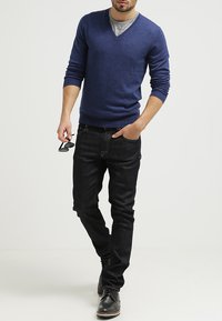 Blauer V-Ausschnitt-Pullover aus leichtem Material, kombiniert mit dunklen Jeans. Das Model trägt schwarze Schuhe und hält in der rechten Hand eine Sonnenbrille.