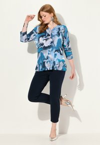 Blå blus med abstrakt mönster och trekvartslånga ärmar, tillsammans med marinblå byxor och silverfärgade högklackade skor. Slät tygtextur.