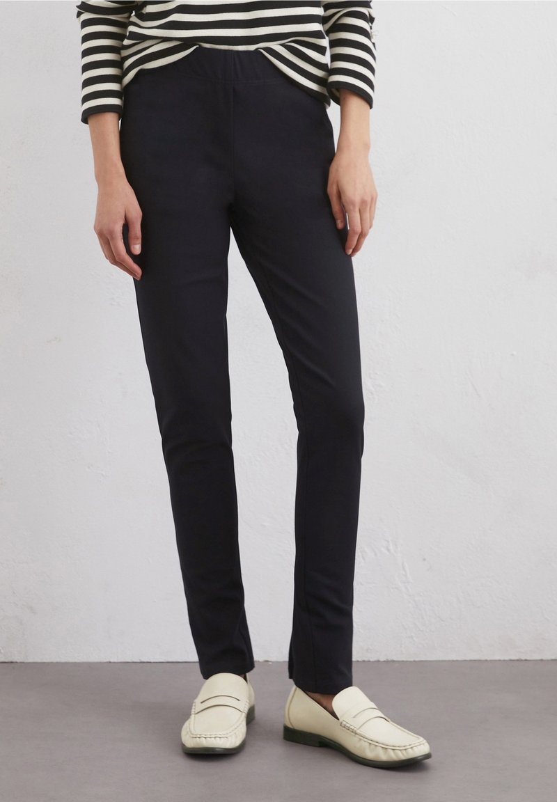 Personne portant un pantalon slim noir, des mocassins crème et un haut manches longues rayé noir et blanc sur un fond clair uni.