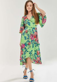Floral knee-length dress in bright green with pink and blue patterns. Features bell sleeves and a v-neck design. Paired with blue strappy heels.