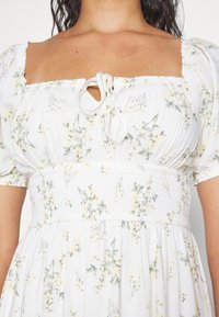 Robe florale blanche avec un décolleté carré, des manches bouffantes courtes et une taille smockée. Présente de délicats motifs floraux jaunes et verts.