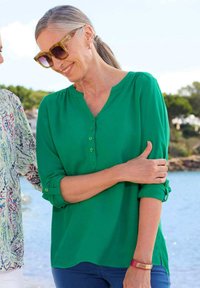 Blusa verde leggera con scollatura a V, dettagli con bottoni e maniche arrotolate. Indossata con jeans blu e un bracciale rosa, in un contesto da spiaggia.