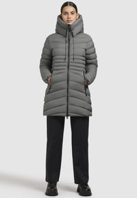 Cappotto imbottito grigio con colletto alto, baffles verticali, chiusura con zip frontale e tasche laterali. Abbinato a pantaloni neri e stivali neri chunky.